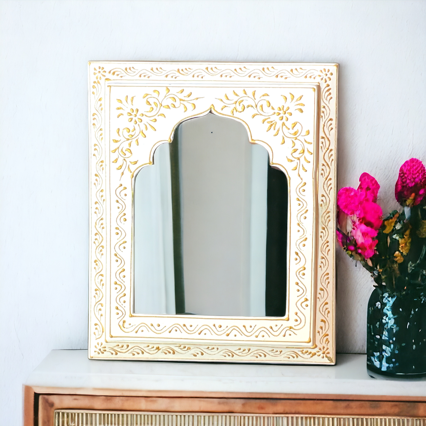 A hand-painted mirror frame with white and golden accents, displayed on a wall above a vase with pink flowers.