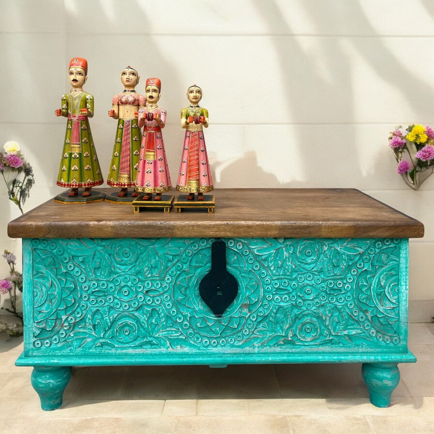 A hand-carved wooden storage trunk with a distressed green finish, featuring black and white detailing, displayed outdoors with a floral arrangement in the background.