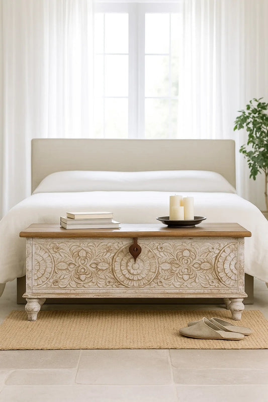 Decorative wooden chest in front of a bed with white bedding and a plant in the corner.
