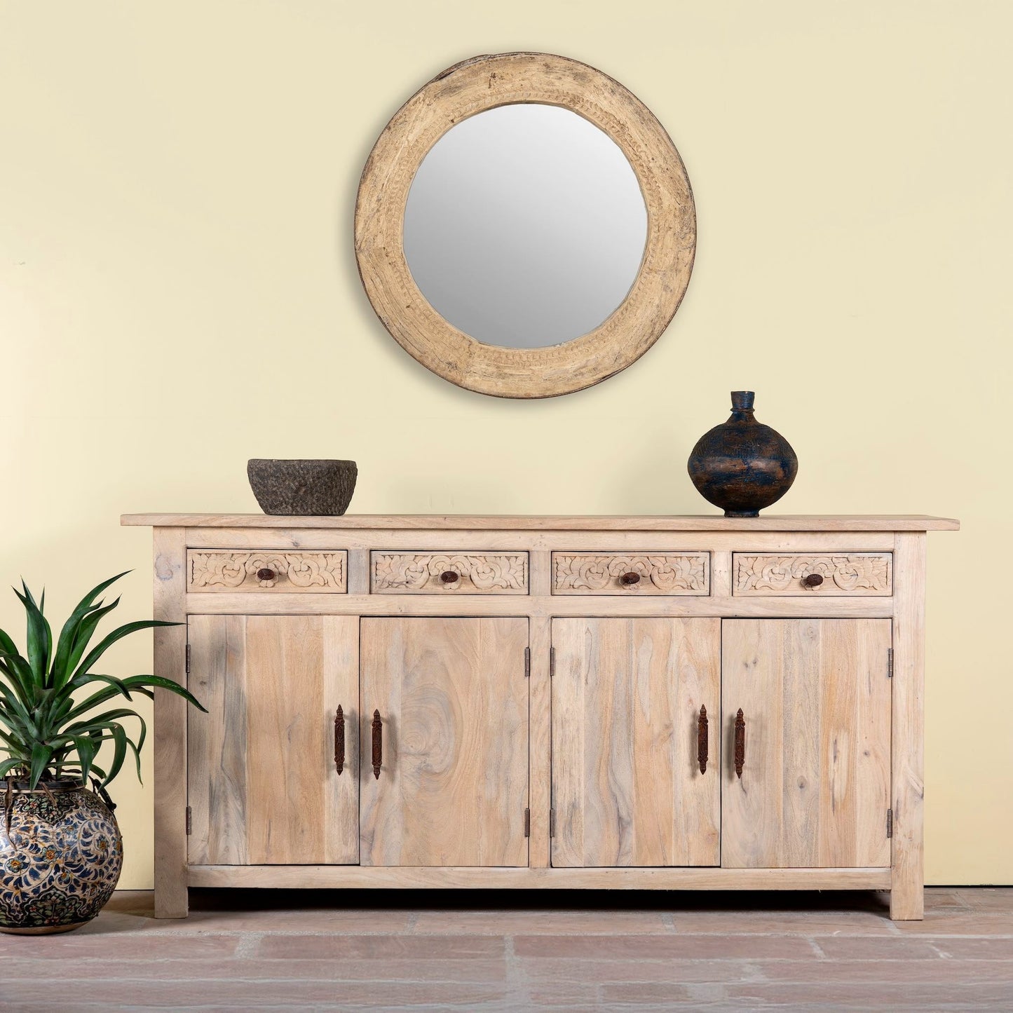 Wooden sideboard with round mirror and vases against a light yellow wall.