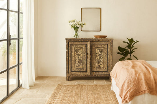 Decorative wooden cabinet with floral arrangement in a room with a bed and large window.