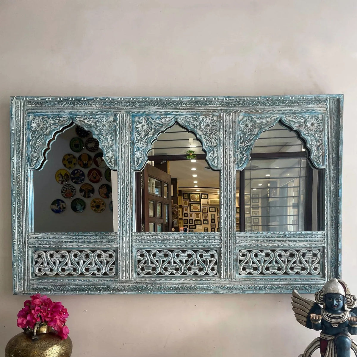 Decorative wall mirror with intricate carvings on a white wall. Large distressed blue handcrafted wooden mirror frame with triple arch Jharokha design and intricate carvings hanging on a wall.Close up of antique teal wood carving on traditional Indian Jharokha mirror frame for wall decor.