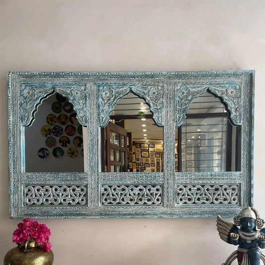 Decorative wall mirror with intricate carvings on a white wall. Large distressed blue handcrafted wooden mirror frame with triple arch Jharokha design and intricate carvings hanging on a wall.Close up of antique teal wood carving on traditional Indian Jharokha mirror frame for wall decor.